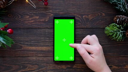 Wall Mural - Women's hands scroll the tape on the phone. Green screen. Top view of smartphone and wooden brown table with christmas decor