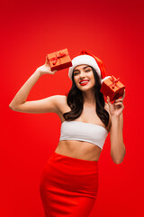 Poster - Sexy woman in santa hat holding gifts and looking at camera isolated on red