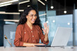 © Liubomir - Successful and beautiful hispanic woman working inside modern office building, businesswoman using laptop for video call smiling and waving, greeting gesture, online conference with colleagues.