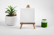 Blank clean white canvas on wood mini easel, mockup on wood tripod, green paint gouache jar and potted plant composition