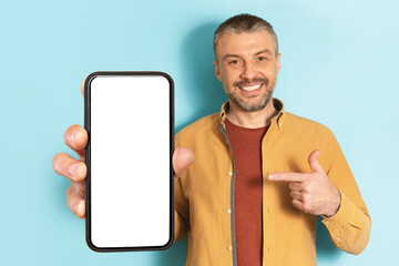Mobile advertisement. Excited middle aged man pointing at cellphone with empty screen on blue background, mockup