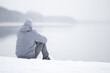 © fotoduets - Young adult man sitting alone on snow at lake shore and looking far away. White cold snowy winter day. Thinking about life. Peaceful atmosphere in nature. Back view.