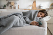© DimaBerlin - Upset sad teenage girl lying under blanket on sofa holding phone, kid spending time staring at screen, sick child using mobile phone while resting in bed at home. Gadget addiction among children