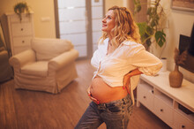Portrait Of Pregnant Woman Standing At Home Holding Hands On Her Belly