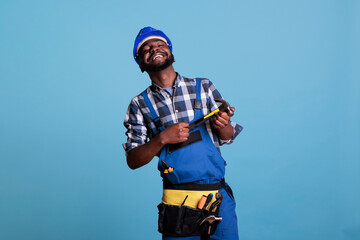 Wall Mural - Studio photo of glad african american builder in helmet and utility belt holding hammer with two hands. Happy construction worker smiling proudly on blue background in studio shot.