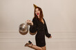 © Look! - Fashionable young caucasian girl in black dress posing with disco ball looking away on white background. Red-haired lady comes off on holiday alone. Party concepts