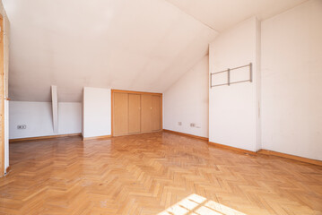 Empty living room with sloping ceilings and wardrobes with wooden sliding doors in the wing, French oak parquet floors