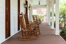 Multiple Vintage Brown Wooden Rocking Chairs On The Veranda Of An Old White Colored Country House With A Wrap Around Porch. There's A Flower Bed And Garden In Front Of The Building With Green Grass. 