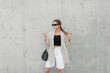 © alones - Stylish beautiful urban model girl with cool sunglasses in fashionable casual clothes with a blazer, top and shorts with a bag near a gray concrete wall in the city