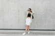 © alones - Fashion beautiful girl with black sunglasses in stylish casual clothes with a blazer, top and shorts with sneakers and a bag near a gray concrete wall