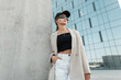 © alones - Pretty urban business woman hipster with cap and fashion sunglasses in blazer, top and shorts stands in modern city. Beauty girl on the street near a concrete wall