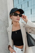 © alones - Street portrait of cool trendy young hipster woman with black cap in fashion casual clothes with blazer, top and bag wears sunglasses in the city. Urban girl