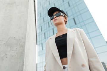 Wall Mural - Beautiful fashionable urban woman with stylish sunglasses and a black cap in fashion casual clothes with a blazer and top walks in the city
