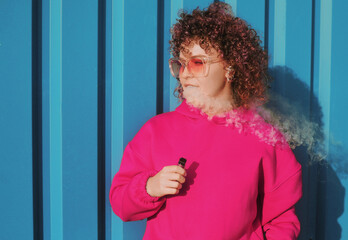 Woman with curly pink hair in magenta hoodie vaping outdoors. Smiling happy woman concept.