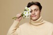 © Tatiana - A handsome man with light white skin, with dark, short, red hair combed back, in a beige turtleneck with a high collar stands on a dark beige background with a bouquet of daisies near his face.Close
