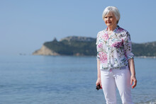 Bella Signora Anziana Passeggia In Riva Al Mare, Sullo Sfondo Si Vede La Spiaggia Le Montagne