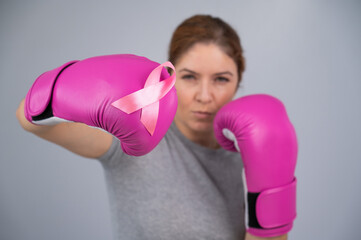 Caucasian woman in pink boxing gloves with a pink ribbon on her chest on a gray background. Fight against breast cancer. 