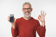 © InsideCreativeHouse - Smiling happy caucasian mature middle-aged man holding cellphone smart phone with mockup screen showing okay gesture isolated in white background