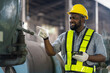 © amorn - African American male worker checking or maintaining machine at the industry factory area. Male worker wear safety helmet and uniform working in the factory