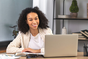 Fototapete - Portrait of a young successful African American businesswoman or an office worker sitting at a desk with a laptop in a modern office, looking at the camera and smiling