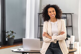 Fototapeta Kuchnia - Young successful African American woman entrepreneur or an office worker stands with crossed arms near a desk in a modern office, looking at the camera and smiling