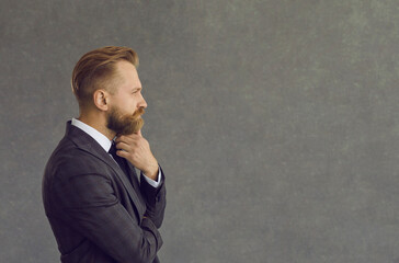 side view portrait handsome bearded man in jacket looking hand on chin at text copyspace on grey bac