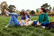 © Xavier Lorenzo - Three happy young women having fun sitting on green grass at park. Multi-ethnic group of female students laughing while relaxing at college campus