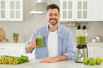 Happy man holding glass of delicious smoothie at white marble table in kitchen