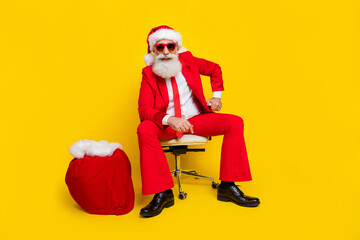 Poster - Photo of positive cheerful retired guy wear red tux claus hat preparing presents sitting chair isolated yellow color background