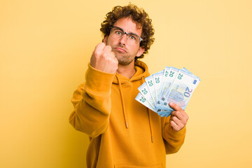 Young curly smart man holding euro banknotes isolated on yellow background showing fist to camera, aggressive facial expression.