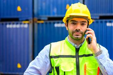 Smiling portrait of handsome male industrial worker foreman in yellow hard hat and safety vest talking on mobile phone controlling goods warehouse at cargo container terminal