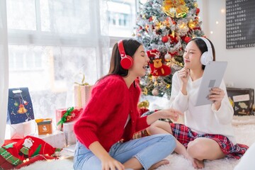 Two young asian female lady friends in a room listening to new songs on their headphone via their digital tablet celebrate Christmas together