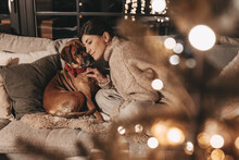 Happy Woman With Dog Cuddling And Unpacking Gifts, Looking Excitedly Into The Box, In A Cozy Environment At Home On The Couch, Mysterious, Warm Colors