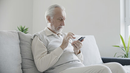 senior man sitting on sofa at home, texting on smartphone, modern technology user