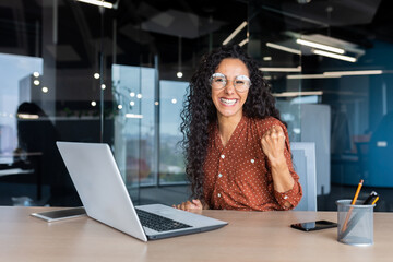 Portrait successful Hispanic business woman office worker with curly hair smiling and looking at camera working at table with laptop holding hand up gesture success and triumph celebrating victory