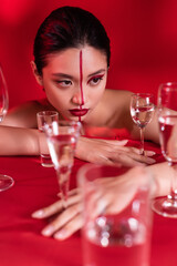 Wall Mural - sensual asian woman with creative visage and naked shoulders posing near glasses of water on red background.