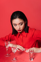 Wall Mural - brunette asian woman in stylish jacket looking away near glasses with pure water on red background.