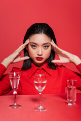 Wall Mural - portrait of asian woman in trendy jacket posing near glasses with clear water isolated on red.