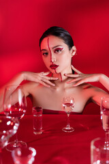 Wall Mural - stylish asian woman with bare shoulders and artistic visage touching neck near various glasses with water on red background.