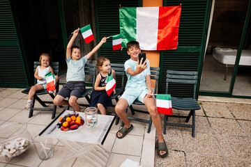 Happy four kids with italian flags celebrating Republic Day of Italy. Boy making selfie on phone.