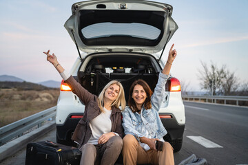 two women mature caucasian female friends sit by the car automobile while parked by the road taking a brake talk in day real people travel vacation copy space happy smile celebrate