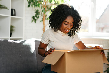 Young african woman sit on couch at home unpacking parcel cardboard box with online purchase