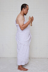 Wall Mural - Side view of Muslim man standing wearing white ihram clothes and praying with an open arm 