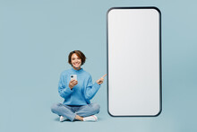 Full Body Young Woman Wear Knitted Sweater Sit Point Finger On Big Huge Blank Screen Mobile Cell Phone With Workspace Mockup Area Use Smartphone Isolated On Plain Pastel Light Blue Cyan Background.