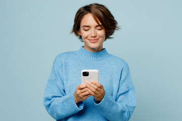 Young smiling happy fun caucasian woman wear knitted sweater hold in hand use mobile cell phone chatting isolated on plain pastel light blue cyan background studio portrait. People lifestyle concept.