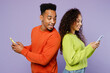 © ViDi Studio - Young fun couple two friends family man woman of African American ethnicity wear casual clothes together hold use mobile cell phone stand back to back peep isolated on plain light purple background.