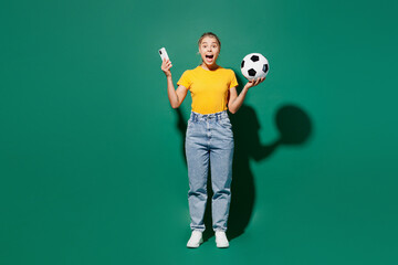 Full body young excited woman fan wears basic yellow t-shirt cheer up support football sport team hold soccer ball watch tv live stream use mobile cell phone isolated on dark green background studio.