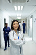 © wavebreak3 - Vertical portrait of smiling asian female doctor in busy hospital corridor, copy space
