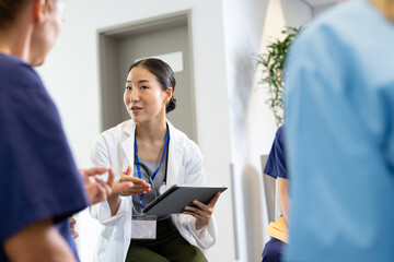 Wall Mural - Smiling asian female doctor with tablet sitting in discussion with diverse colleagues at hospital