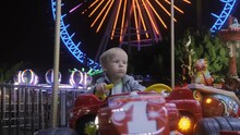 A Cute Little Boy Is Driving While Sitting On A Racing Car On An Amusement Park Ride In The Evening Against The Background Of A Ferris Wheel. High Quality 4k Footage